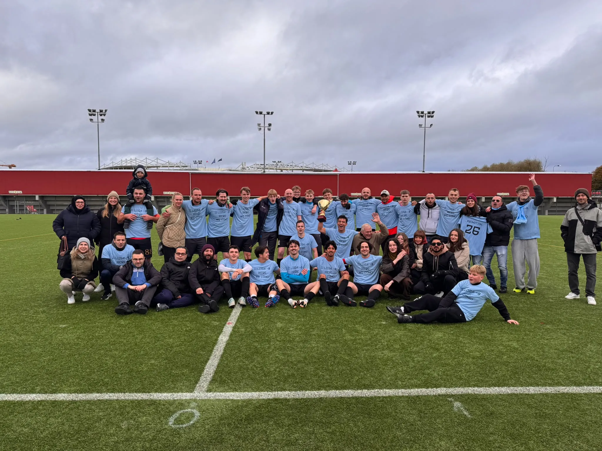 Rumori players, staff, families and friends on the pitch after winning III liiga North/West in 2026, in shirts that read “II liiga B, here we come”, with the trophy